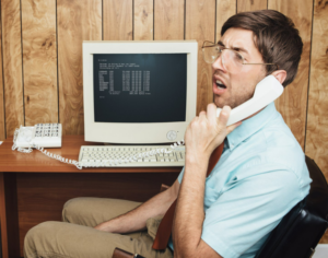 a man sitting on a black office chair on a phone call using landline phone with a white computer on a brown desk showing the power of embracing computers 